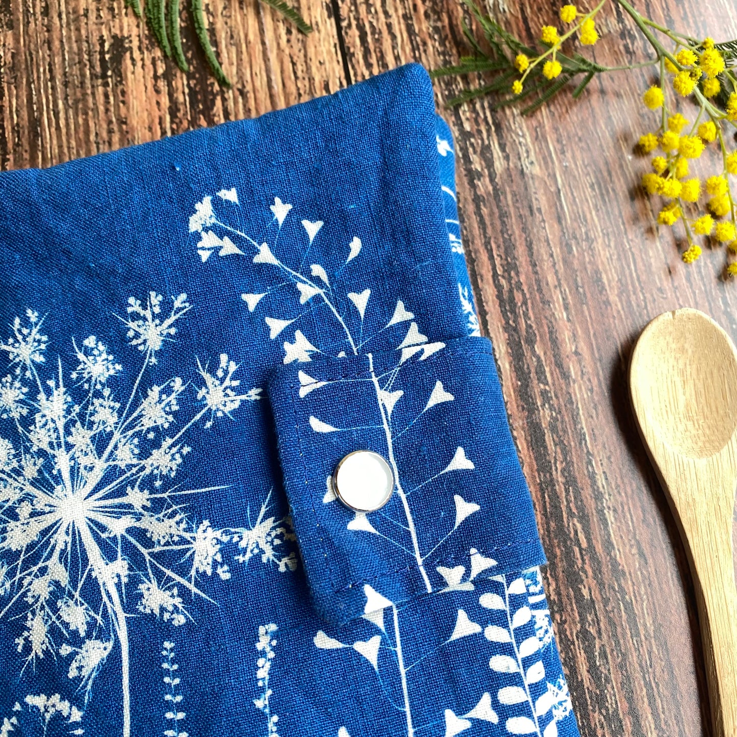 Détail de pochette en tissu cyanotype, motif fleur bleue, bouton nacré. Posée sur table bois, à coté d'une cuillère.
