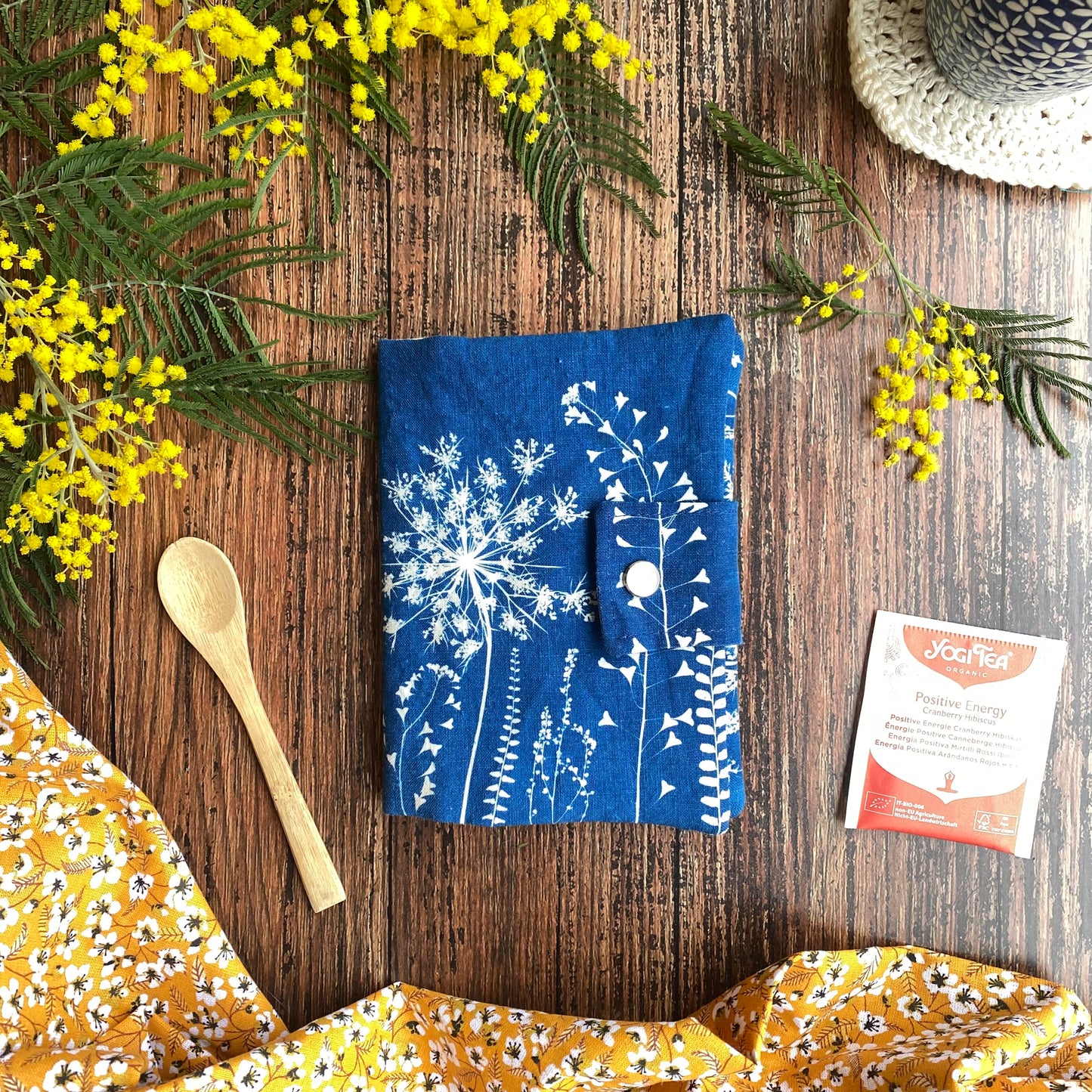 Pochette bleue en tissu cyanotype, pour ranger les sachets de thé. Table en bois, fleur de mimosa. Tasse sur napperon.