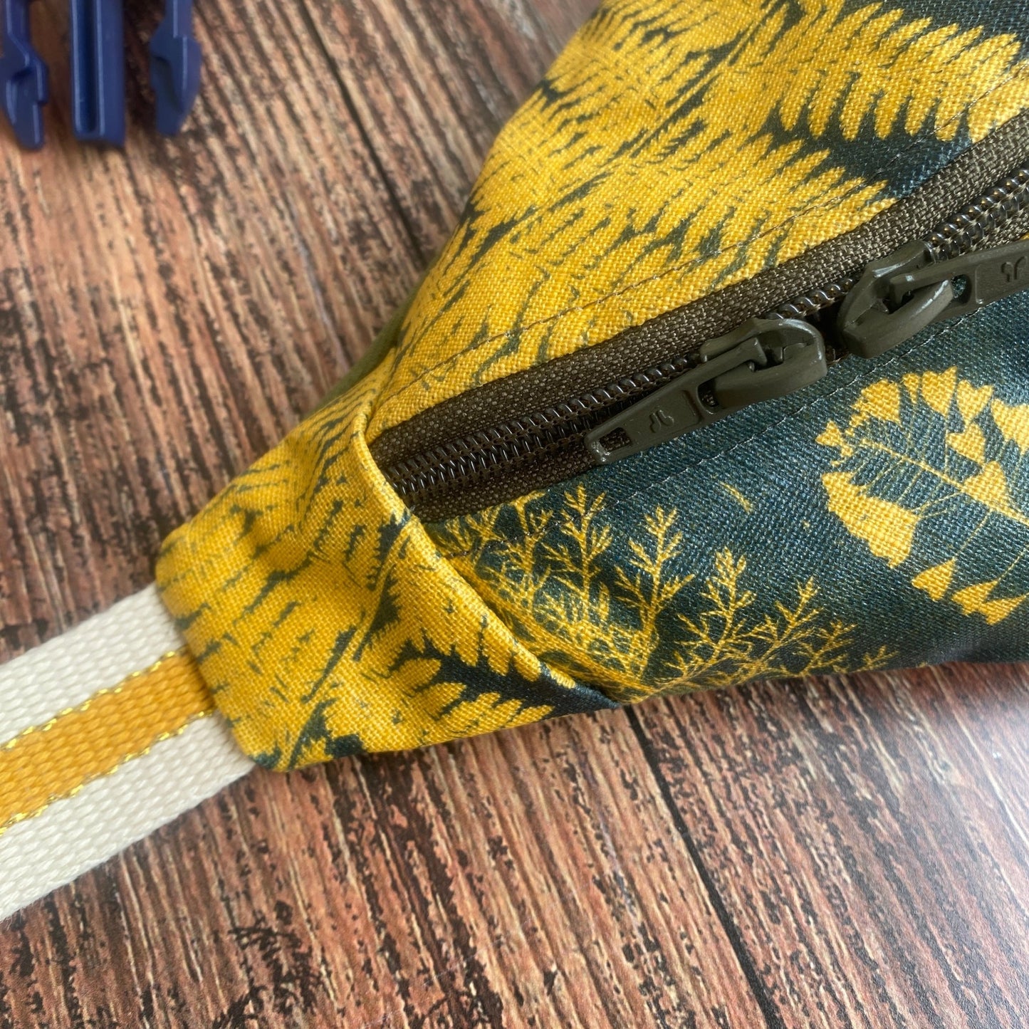 Sac banane en tissu fleuri kaki, format XL. Détail de la sangle beige et jaune avec un liseré doré.