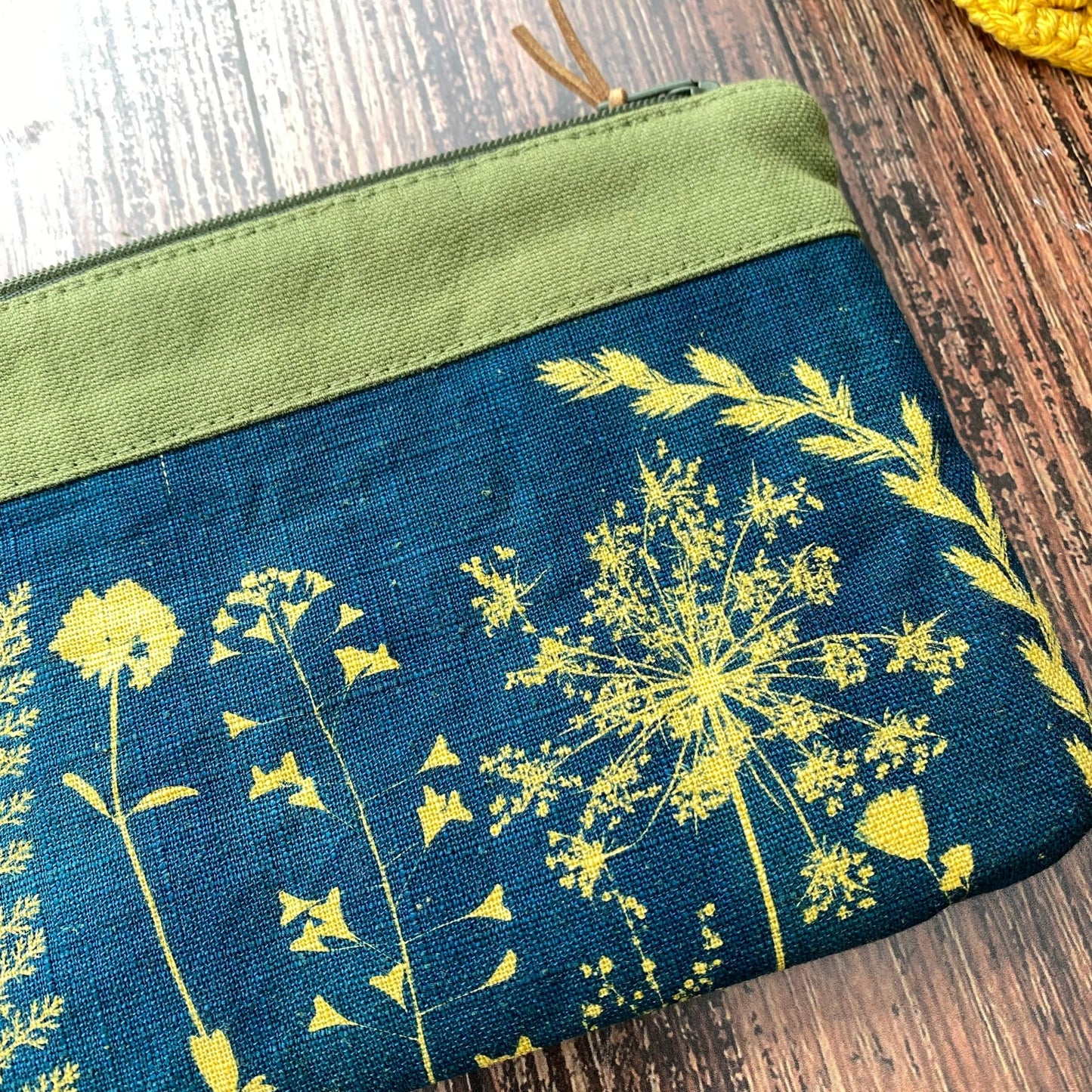 Détail d'une pochette zipée fait main ornée d'un cyanotype de fleurs sur tissu kaki posée sur une table en bois.