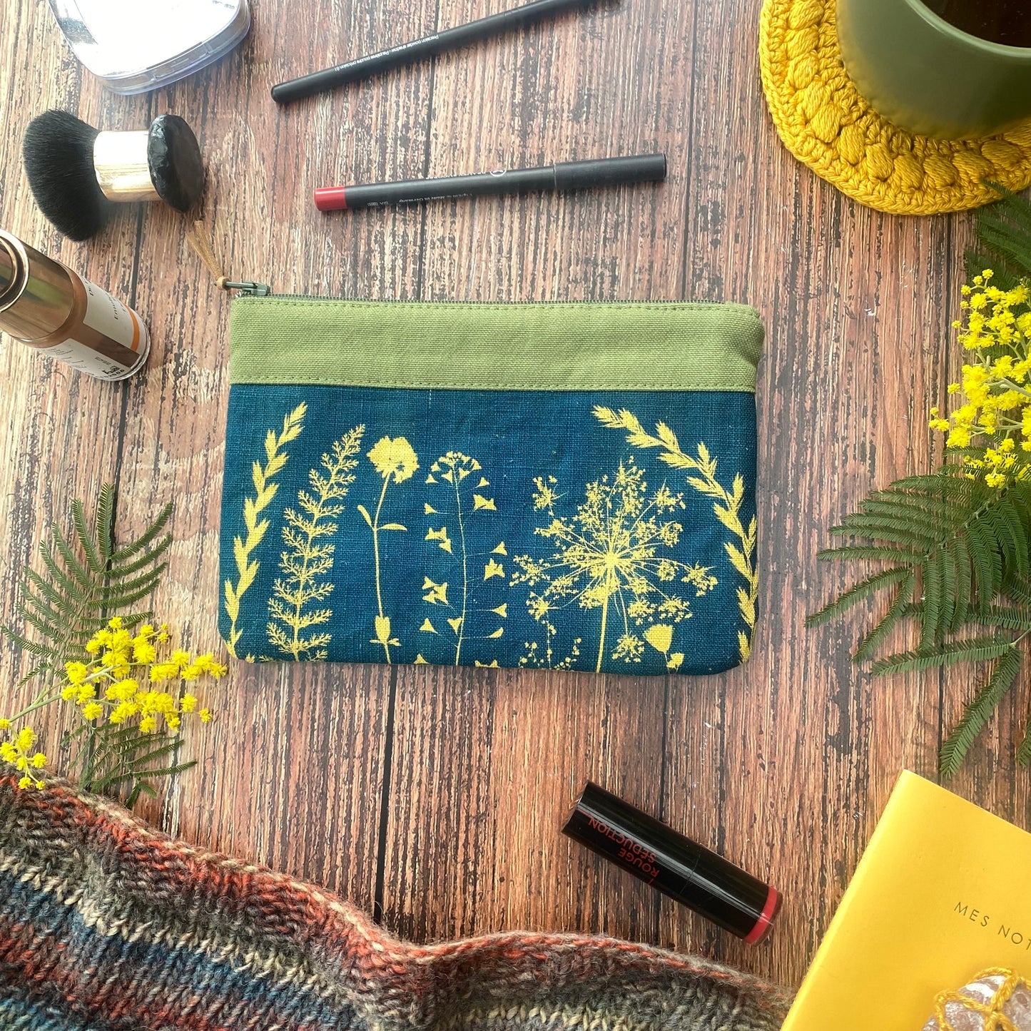 Pochette tissu cyanotype kaki, bicolore, sur table bois, accessoires maquillage, fleurs, tasse, carnet et tricot.