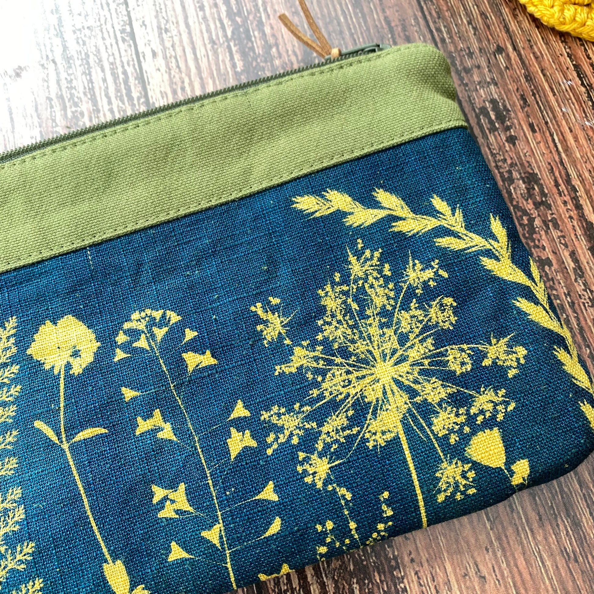 Détail d'une pochette zipée fait main ornée d'un cyanotype de fleurs sur tissu kaki posée sur une table en bois.
